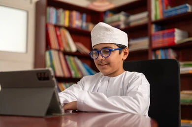 Reviewing daily lessons, the new semester, an Omani Gulf Arab student wearing a dishdasha and a kumma sits in the school library solving assignments and attending online school lectures via a tablet. Reviewing daily lessons, the new semester, an Omani Gulf Arab student wearing a dishdasha and a kumma sits in the school library solving assignments and attending online school lectures via a tablet.