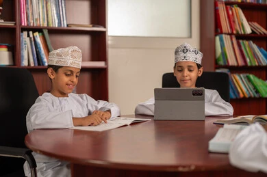 Solving class assignments, gestures of integration and focus, the concept of diligence and the pursuit of excellence, the importance of knowledge in developing mental abilities, education in the Sultanate of Oman, two Arab Gulf Omani students wearing the dishdasha and kumma sitting inside the school library, using tablets in studying.