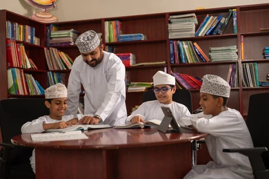 Enjoying reading school books, using a tablet for learning and entertainment, an Arab Gulf Omani teacher helping his students, Arab Gulf Omani students wearing the dishdasha and kumma sitting in the school library with their teacher, doing homework, education in the Sultanate of Oman.