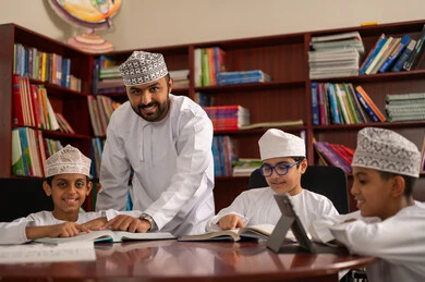 استخدام جهاز التابلت في التعلم والترفيه، معلم عربي خليجي عماني يقوم بمساعده طلابه، طلاب عربيين خليجيين عمانيين يرتدون الدشداشة و الكمة يجلسون في المكتبة المدرسية برفقة معلمهم ، الاستمتاع بقراءة الكتب المدرسية، تأدية الواجبات المدرسية، التعليم في سلطنه عمان