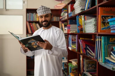 قراءة الكتب المفيدة، مفهوم المطالعة والثقافة ، التعليم في سلطنة عمان، ممارسة الهوايات المفضلة ، معلم عربي خليجي عماني يرتدي الدشداشة و الكمة يقف في المكتبة المدرسية يمسك بيدية كتاب