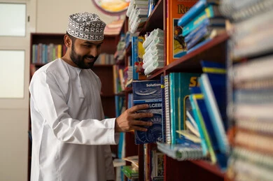 Acquiring knowledge through reading, the concept of reading and culture, education in the Sultanate of Oman, practicing favorite hobbies, an Arab Gulf Omani teacher wearing a dishdasha and kumma stands in the school library holding a book in his hands.