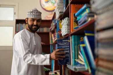 Reading purposeful and informative books, the concept of reading and culture, education in the Sultanate of Oman, practicing favorite hobbies, an Arab Gulf Omani teacher wearing a dishdasha and a kumma stands in the school library holding a book in his hands.