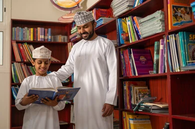 تقوية المهارات القرائية للطلبة، حب الثقافة والاستطلاع، زيارة مكتبة المدرسة، طالب عربي خليجي عماني يرتدي الدشداشة و الكمة يقف برفقة معلمه يحمل بيده كتاب، التعليم والدراسة في سلطنه عمان، قضاء وقت الفراغ بالتعلم والتثقف الذاتي