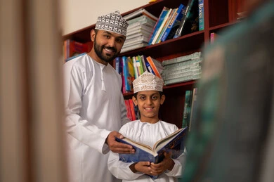 تنمية مهارة القراءة للطلاب، حب الثقافة والاستطلاع، زيارة مكتبة المدرسة، طالب عربي خليجي عماني يرتدي الدشداشة و الكمة يقف برفقة معلمه يحمل بيده كتاب، التعليم والدراسة في سلطنه عمان، قضاء وقت الفراغ بالتعلم والتثقف الذاتي