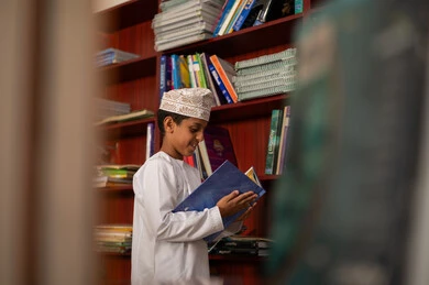 زيارة مكتبة المدرسة، حب الثقافة والاستطلاع، بيئة تعليمية متطورة، تأدية الواجبات المدرسية ، طالب عربي خليجي عماني يرتدي الدشداشة و الكمة يقف امام مكتبة خشبية يمسك بيده كتاب، الاستمتاع بقراءة الكتب المدرسية