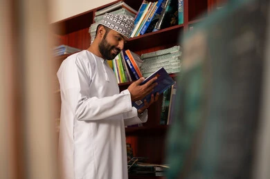 التعمق في الكتب الثرية بالمعلومات، مفهوم المطالعة والثقافة ، التعليم في سلطنة عمان، ممارسة الهوايات المفضلة ، معلم عربي خليجي عماني يرتدي الدشداشة و الكمة يقف بجانب مكتبة خشبية يمسك بيدية كتاب