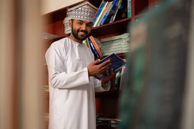 Reading useful books, the concept of reading and culture, education in the Sultanate of Oman, practicing favorite hobbies, an Arab Gulf Omani teacher wearing a dishdasha and a kumma stands next to a wooden library holding a book in his hands.