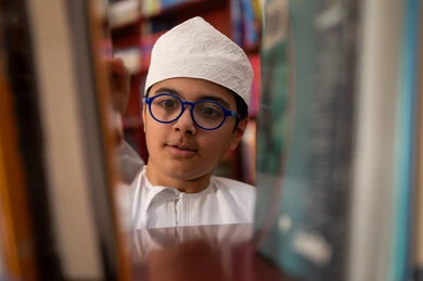 الاهتمام بالمطالعة وتطويرها، بيئه مدرسيه تعليميه متطوره، بناء مستقبل مشرق للمجتمع، مفهوم القراءه والثقافه والاستطلاع، صورة مقربة لطالب عربي خليجي عماني يرتدي الدشداشة و الكمة يقف بجوار مكتبة خشبية فى المدرسة , مدارس أكاديميه في سلطنه عمان