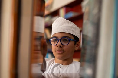 حب القراءة وممارستها، بيئه مدرسيه تعليميه متطوره، بناء مستقبل مشرق للمجتمع، مفهوم القراءه والثقافه والاستطلاع، صورة مقربة لطالب عربي خليجي عماني يرتدي الدشداشة و الكمة يقف بجوار مكتبة خشبية فى المدرسة , مدارس أكاديميه في سلطنه عمان