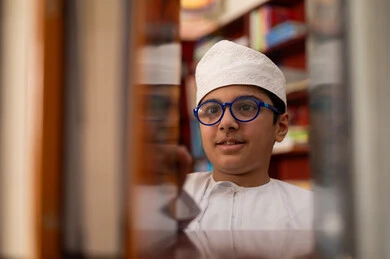 قضاء الوقت في المطالعة، بيئه مدرسيه تعليميه متطوره، بناء مستقبل مشرق للمجتمع، مفهوم القراءه والثقافه والاستطلاع، صورة مقربة لطالب عربي خليجي عماني يرتدي الدشداشة و الكمة يقف بجوار مكتبة خشبية فى المدرسة , مدارس أكاديميه في سلطنه عمان