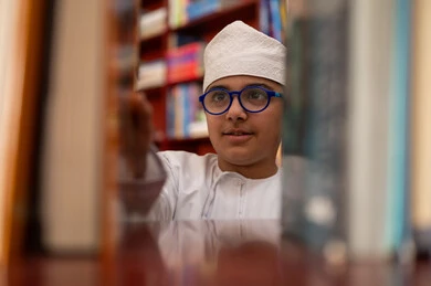 حب قراءة الكتب والمطالعة، بيئه مدرسيه تعليميه متطوره، بناء مستقبل مشرق للمجتمع، مفهوم القراءه والثقافه والاستطلاع، صورة مقربة لطالب عربي خليجي عماني يرتدي الدشداشة و الكمة يقف بجوار مكتبة خشبية فى المدرسة , مدارس أكاديميه في سلطنه عمان