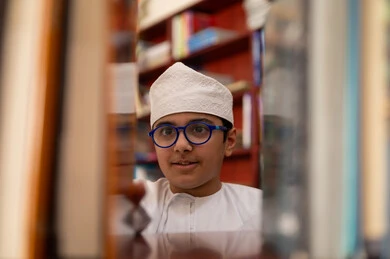 Enjoying reading time, a developed educational school environment, building a bright future for the community, the concept of reading, culture, and exploration, a close-up image of an Arab Gulf Omani student wearing a dishdasha and kumma standing next to a wooden library in the school, academic schools in the Sultanate of Oman.