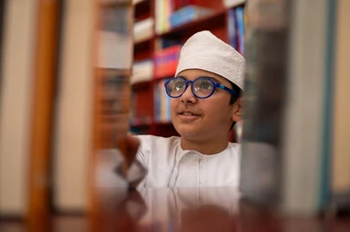 الاستمتاع بقراءة الكتب المدرسية، ممارسة هوايه المطالعة، بيئه مدرسيه تعليميه متطوره، بناء مستقبل مشرق للمجتمع، مفهوم القراءه والثقافه والاستطلاع، صورة مقربة لطالب عربي خليجي عماني يرتدي الدشداشة و الكمة يقف بجوار مكتبة خشبية فى المدرسة , مدارس أكاديميه في سلطنه عمان