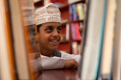 Practicing the hobby of reading, a developed educational school environment, building a bright future for the community, the concept of reading, culture, and exploration, a close-up image of an Arab Gulf Omani student wearing a dishdasha and kumma standing next to a wooden library in the school, academic schools in the Sultanate of Oman, enjoying reading school books.