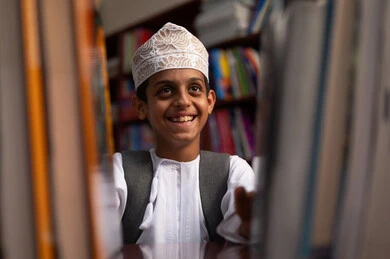A developed educational school environment, building a bright future for the community, the concept of reading, culture, and exploration, a close-up image of an Arab Gulf Omani student wearing a dishdasha and kumma standing next to a wooden library in the school, academic schools in the Sultanate of Oman, practicing the hobby of reading, enjoying reading school books.