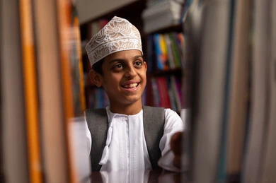 بناء مستقبل مشرق للمجتمع، مفهوم القراءه والثقافه والاستطلاع، صورة مقربة لطالب عربي خليجي عماني يرتدي الدشداشة و الكمة يقف بجوار مكتبة خشبية فى المدرسة , مدارس أكاديميه في سلطنه عمان، بيئه مدرسيه تعليميه متطوره ، ممارسة هوايه المطالعة، الاستمتاع بقراءة الكتب المدرسية