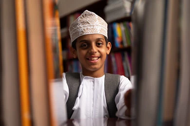 مفهوم القراءه والثقافه والاستطلاع، صورة مقربة لطالب عربي خليجي عماني يرتدي الدشداشة و الكمة يقف بجوار مكتبة خشبية فى المدرسة , مدارس أكاديميه في سلطنه عمان، بناء مستقبل مشرق للمجتمع، بيئه مدرسيه تعليميه متطوره ، ممارسة هوايه المطالعة، الاستمتاع بقراءة الكتب المدرسية