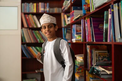 Looking at the camera with expressions of happiness and joy, academic schools in the Sultanate of Oman, practicing favorite hobbies, an Arab Gulf Omani student wearing a dishdasha and kumma and a school bag standing next to a wooden library in the school library.