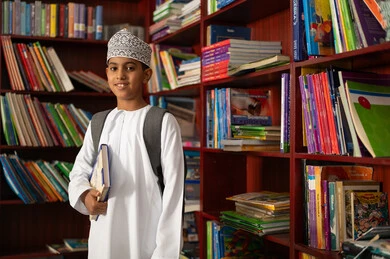 Education in the Sultanate of Oman, practicing favorite hobbies, an Arab Gulf Omani student wearing a dishdasha and kumma and carrying a school bag stands next to a wooden bookshelf in the school library, enjoying reading school books.