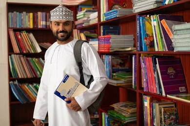 التربية والتعليم بالمناهج المقررة للتلاميذ، جودة التعليم في سلطنه عمان، معلم عربي خليجي عماني يرتدي الدشداشة و الكمة يحمل حقيبة مدرسية على ظهره يقف داخل المكتبة المدرسية ، النظر الى الكاميرا بتعابير السعادة و السرور