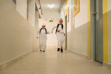 تقوية الروابط الاجتماعية بين الطلاب، الإستعداد للفصل الدراسي الجديد، وقت اللعب والترفيه ، طالبان عربيان خليجيان يرتديان الدشداشة و الكمة و الحقيبة المدرسية يسيران داخل مبني المدرسه, ايماءات السعاده والمرح, مدارس أكاديميه في سلطنة عمان