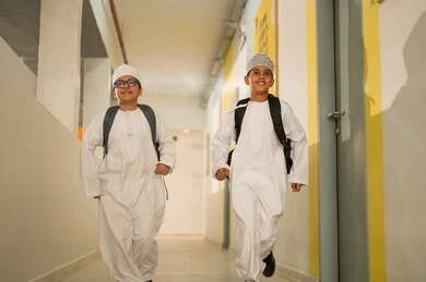 Enhancing friendly communication among students, preparing for the new school semester, play and leisure time, two Gulf Arab students wearing the dishdasha and the kumma and carrying school bags walking inside the school building, gestures of happiness and fun, academic schools in the Sultanate of Oman.