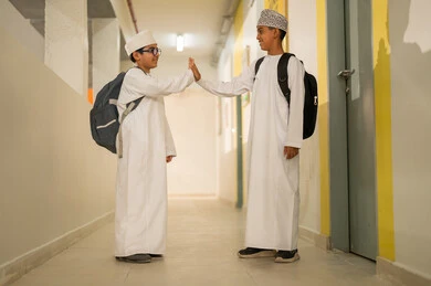 Building relationships and forming new friendships, welcoming and greeting, preparing for the new school semester, play and entertainment time, two Gulf Arab students wearing the dishdasha and the kumma and carrying school bags stand in the school hallway, high-fiving each other, gestures of happiness and fun, academic schools in the Sultanate of Oman.
