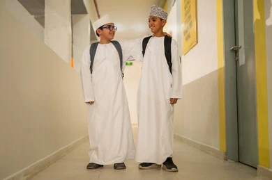 دعم العلاقات الودية بين الطلبة، الإستعداد للفصل الدراسي الجديد، وقت اللعب والترفيه ، طالبان عربيان خليجيان يرتديان الدشداشة و الكمة و الحقيبة المدرسية يخرجان الى بهو المدرسه, ايماءات السعاده والمرح, مدارس أكاديميه في سلطنة عمان