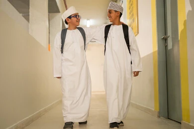 Fostering a spirit of camaraderie within the student community, preparing for the new school semester, play and leisure time, two Gulf Arab students wearing the dishdasha and the kumma and carrying school bags are heading to the school hall, gestures of happiness and fun, academic schools in the Sultanate of Oman.