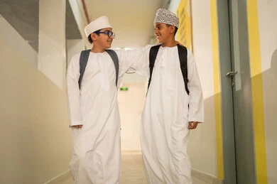 تقوية علاقات المحبة بين الطلاب، الإستعداد للفصل الدراسي الجديد، وقت اللعب والترفيه ، طالبان عربيان خليجيان يرتديان الدشداشة و الكمة و الحقيبة المدرسية يخرجان الى بهو المدرسه, ايماءات السعاده والمرح, مدارس أكاديميه في سلطنة عمان