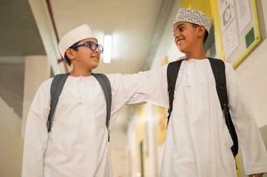 مدارس أكاديميه في سلطنة عمان، الإستعداد للفصل الدراسي الجديد، مفهوم الموده والالفه، وقت اللعب والترفيه ، طالبان عربيان خليجيان يرتديان الدشداشة و الكمة و الحقيبة المدرسية يخرجان الى بهو المدرسه, ايماءات السعاده والمرح