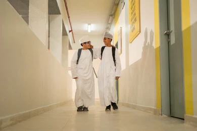 الإستعداد للفصل الدراسي الجديد، مفهوم الموده والالفه، وقت اللعب والترفيه ، طالبان عربيان خليجيان يرتديان الدشداشة و الكمة و الحقيبة المدرسية يخرجان الى بهو المدرسه, ايماءات السعاده والمرح, مدارس أكاديميه في سلطنة عمان