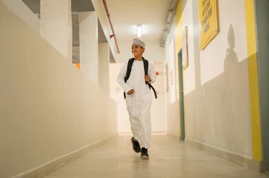 Determination and hard work to achieve excellence, gestures of happiness and joy, the back-to-school season, an Arab Gulf Omani student wearing a dishdasha and kumma carrying a school backpack walking in the school hallway.