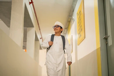 Diligence and ambition to achieve excellence, gestures of happiness and joy, the back-to-school season, an Arab Gulf Omani student wearing a dishdasha and kumma carrying a school backpack walking in the school hallway.