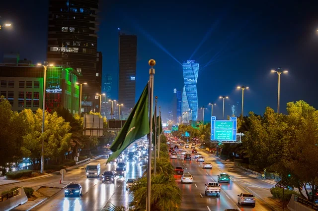The flag of the Kingdom is the green banner of monotheism, blue road signs, the architectural engineering art of a lit tower in the city of Riyadh at night, famous commercial, economic, and tourist landmarks in the capital, the architectural engineering art of the facilities, and urban growth and development in the modern and advanced style of towers.