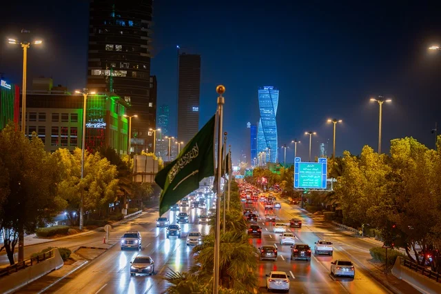 Blue road signs, the Kingdom's flag, the green banner of unification, the architectural engineering art of a lit tower in Riyadh at night, famous commercial, economic, and tourist landmarks in the capital, the architectural engineering art of facilities, urban growth and development in the modern and advanced style of towers.