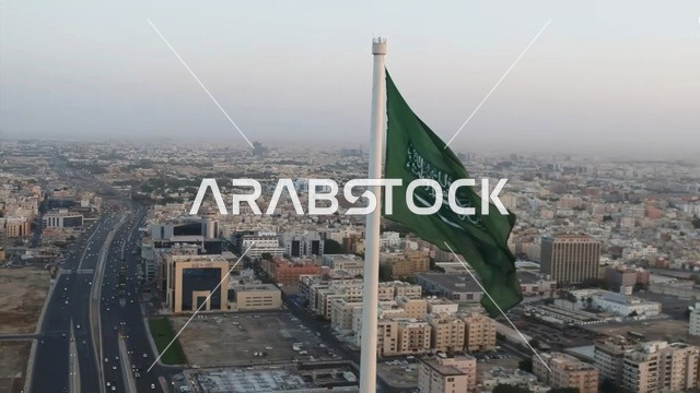 رمز المملكة العربية السعودية، سارية علم السعودية ترفرف فوق المباني السكنية في السماء وقت غروب الشمس، الاحتفال بيوم العلم السعودي 11 مارس، راية التوحيد والإسلام وشعار الهوية الوطنية، ذكرى اليوم الوطني 23 سبتمبر، خلفية السماء زرقاء