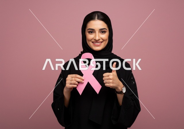 حملة توعية للكشف المبكر عن سرطان الثدي،رمز الحملة العالمية، اليوم العالمي للتوعية والكشف المبكر، تحدي الأمراض الخبيثة، بورتريه مقرب لإمرأة عربية خليجي سعودية ترتدي الحجاب والعباءة السوداء وتحمل الشريط الوردي، النظر الى الكاميرا بايماءات الجودة،  خلفية وردية