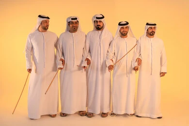 الحفاظ على التراث الشعبي في المناسبات الوطنية والاجتماعية، بورتريه لمجموعة من الرجال العرب الخليجيين الإماراتيين يرتدون الكندورة والغترة البيضاء يؤدون رقصة العيالة الشعبية بعصا الخيزران، أجواء تراثية تعبّر عن الفخر بالموروث الثقافي الإماراتي، إيقاعات متناغمة وحركات جماعية تعكس روح الوحدة والاعتزاز بالهوية الوطنية