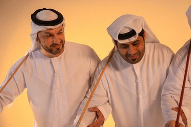 مجموعة من الرجال العرب الخليجيين الإماراتيين يرتدون الكندورة والغترة البيضاء يؤدون رقصة العيالة الشعبية بعصا الخيزران، أجواء تراثية تعبّر عن الفخر بالموروث الثقافي الإماراتي،إيقاعات متناغمة وحركات جماعية تعكس روح الوحدة والاعتزاز بالهوية الوطنية، الحفاظ على التراث الشعبي في المناسبات الوطنية والاجتماعية.
