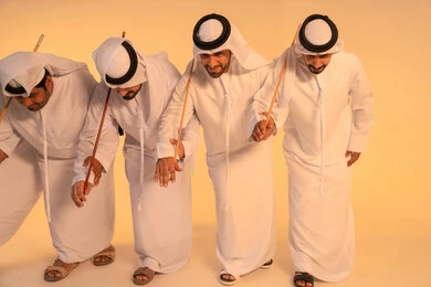 مجموعة من الرجال العرب الخليجيين الإماراتيين يرتدون الكندورة والغترة البيضاء يؤدون رقصة العيالة الشعبية بعصا الخيزران، أجواء تراثية تعبّر عن الفخر بالموروث الثقافي الإماراتي،إيقاعات متناغمة وحركات جماعية تعكس روح الوحدة والاعتزاز بالهوية الوطنية، الحفاظ على التراث الشعبي في المناسبات الوطنية والاجتماعية.