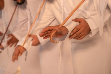 Heritage atmosphere expressing pride in the Emirati cultural heritage, harmonious rhythms and collective movements reflecting the spirit of unity and pride in national identity, a group of Arab Gulf Emirati men wearing the kandura and white ghutrah performing the traditional Ayala dance with bamboo sticks, preserving the folk heritage during national and social occasions.