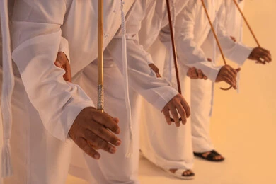 National celebration and belonging, harmonious rhythms and collective movements reflect the spirit of unity and pride in national identity. A group of Emirati Gulf Arab men wearing the kandura and white ghutrah perform the traditional Ayala dance with bamboo sticks. The atmosphere is cultural, expressing pride in the Emirati cultural heritage and preserving the folk heritage during national and social occasions.