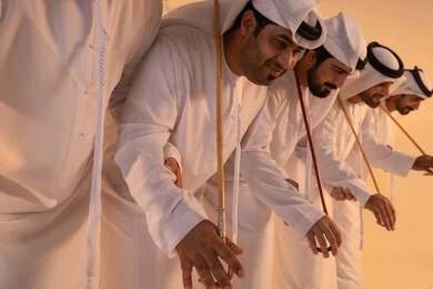 Harmonious rhythms and collective movements reflect the spirit of unity and pride in national identity. A group of Emirati Gulf Arab men wearing the kandura and white ghutrah perform the traditional Ayala dance with bamboo sticks. The atmosphere is cultural, expressing pride in the Emirati cultural heritage and preserving the folk heritage during national and social occasions.