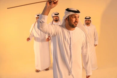 Embodiment of pride in Emirati customs and traditions, an Arab Gulf Emirati man wearing the kandura and ghutrah performs the Ayala dance with a bamboo stick, in a joyful festive atmosphere, showcasing courage, generosity, and national belonging, preserving the popular heritage and cultural identity during national and social occasions.