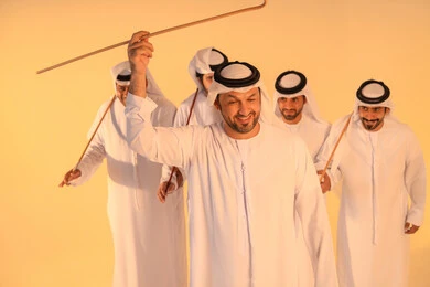 Harmonious rhythms and collective movements reflect the spirit of unity and pride in national identity. A group of Emirati Gulf Arab men wearing the kandura and white ghutrah perform the traditional Ayala dance with bamboo sticks. The atmosphere is cultural, expressing pride in the Emirati cultural heritage and preserving the folk heritage during national and social occasions.