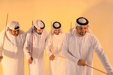 Pride in Emirati customs and traditions, harmonious rhythms and collective movements reflect the spirit of unity and pride in national identity. A group of Emirati Gulf Arab men wearing the kandura and white ghutrah perform the traditional Ayala dance with bamboo sticks, preserving the folk heritage during national and social occasions.