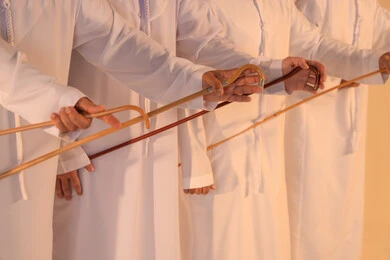 Heritage atmosphere expressing pride in the Emirati cultural heritage, harmonious rhythms and collective movements reflecting the spirit of unity and pride in national identity, a group of Arab Gulf Emirati men wearing the kandura and white ghutrah performing the traditional Ayala dance with bamboo sticks, preserving the folk heritage during national and social occasions.