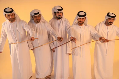 الحفاظ على التراث الشعبي في المناسبات الوطنية والاجتماعية ،إيقاعات متناغمة وحركات جماعية تعكس روح الوحدة والاعتزاز بالهوية الوطنية،مجموعة من الرجال العرب الخليجيين الإماراتيين يرتدون الكندورة والغترة البيضاء يؤدون رقصة العيالة الشعبية بعصا الخيزران، أجواء تراثية تعبّر عن الفخر بالموروث الثقافي الإماراتي، الحفاظ على التراث الشعبي في المناسبات الوطنية والاجتماعية.